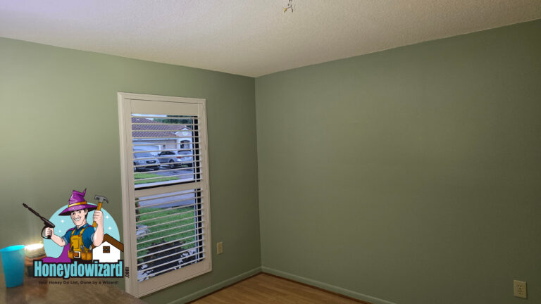 A small, empty room with light green walls, wood floors, a window with white blinds, and an overhead light. The Honeydowizard logo appears in the lower left corner. Honey Do List Wizard Handyman
