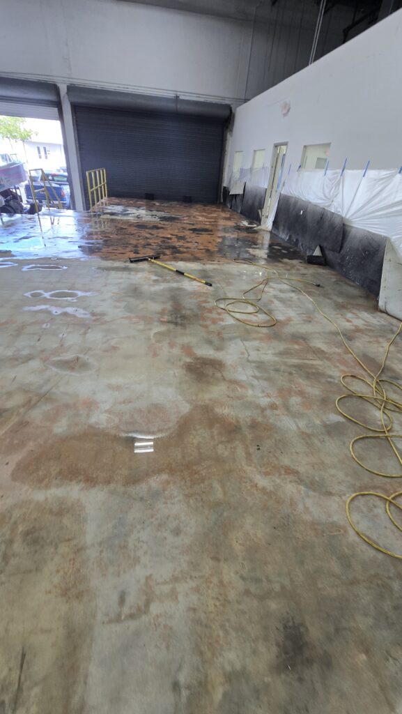 A large indoor space with a concrete floor, partially wet and stained with rust-colored patches—evidence of recent pressure washing. Cleaning equipment and yellow cables are strewn across the floor near a partially open garage door in Broward County. Honey Do List Wizard Handyman