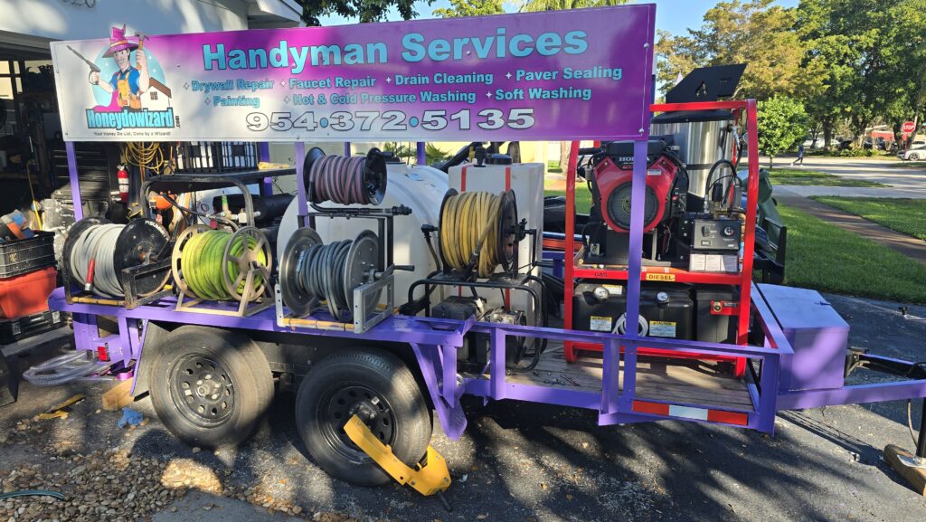 A purple and pink Honey Do Wizard trailer is parked outside, loaded with equipment, hoses, and tanks. The sign lists services like drywall repair, Commercial Hot Water Pressure Washing, drain cleaning, and paver sealing with a phone number displayed prominently. Honey Do List Wizard Handyman